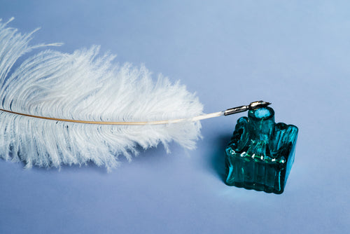 white feather quill resting on an ink bottle on blue background