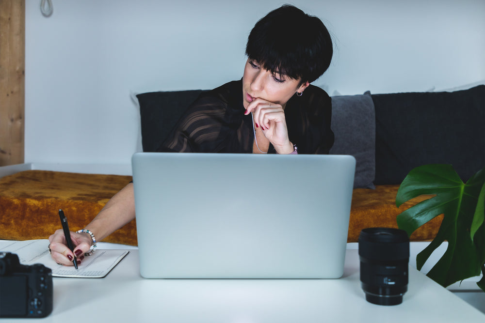 woman at laptop in thought as she writes in a notebook