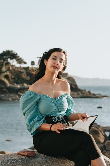 woman holds a book and sits on rocks by the water