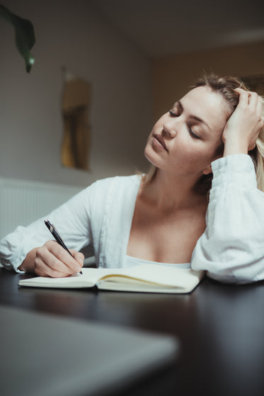 woman holds her head thinking while writing in a journal
