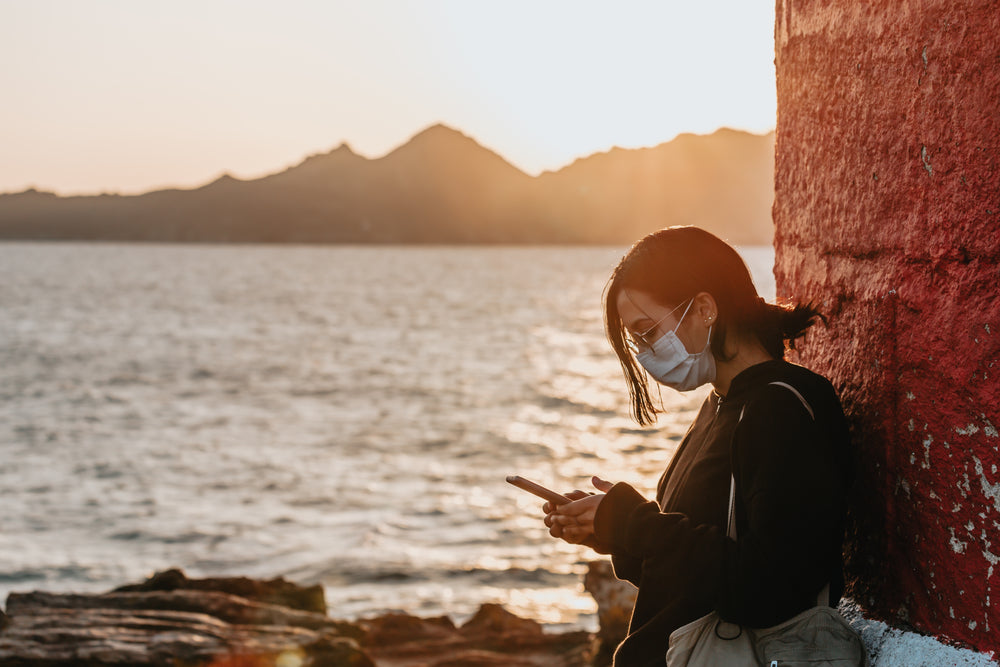 woman in a facemask looks at her phone standing by water