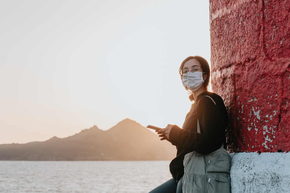 woman in facemask leans against a wall by water