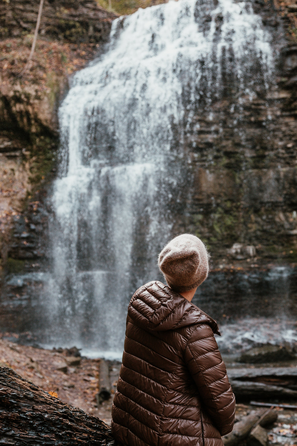 woman in winter fashion sits by foot of waterfall