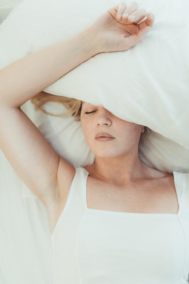 woman lays in a bed with a pillow over her head