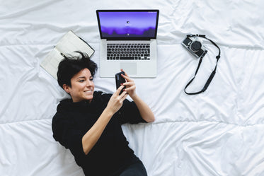 woman lays on bed looking at her cell phone with a laptop nearby