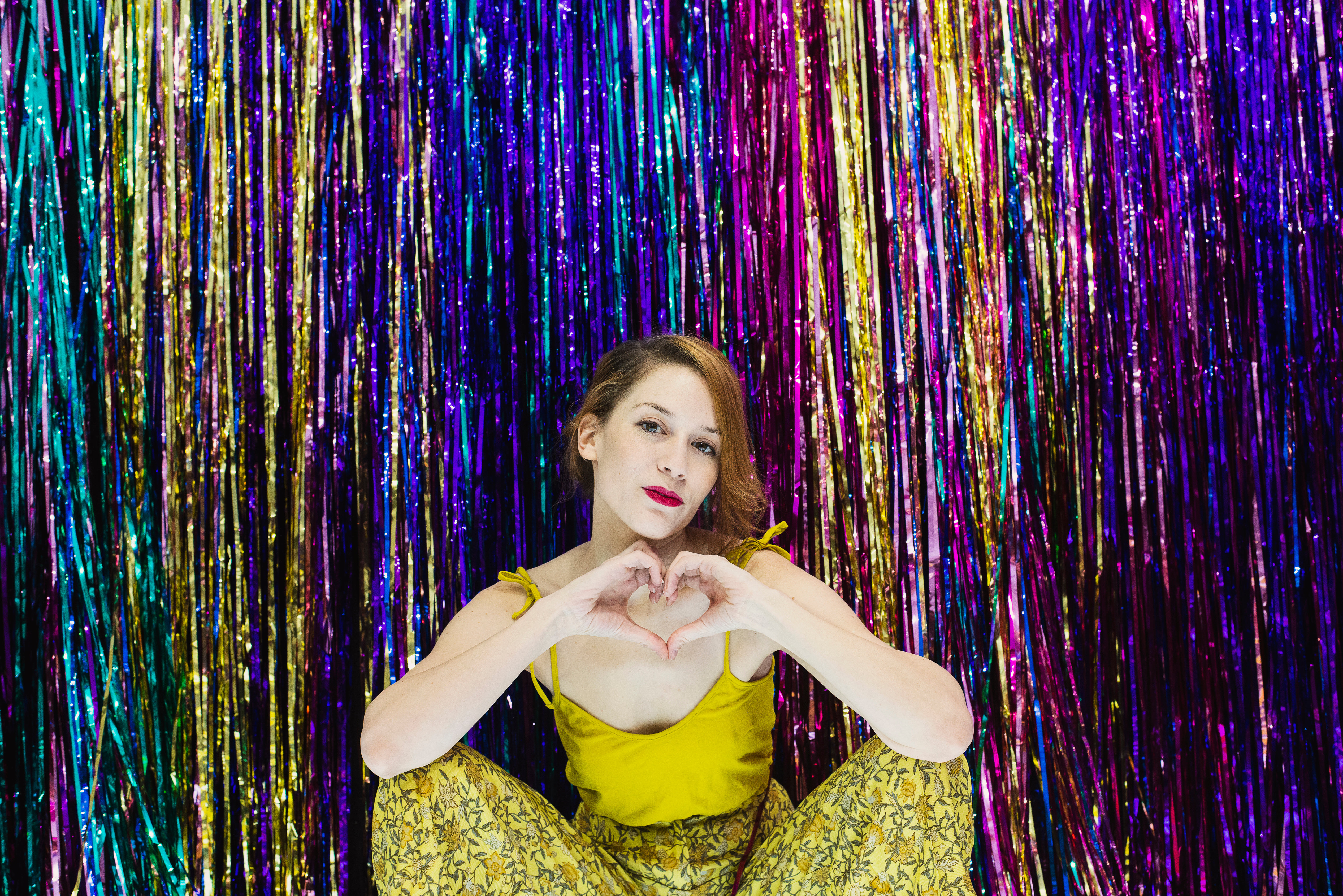 Woman Makes Hand Heart Gesture In Front Of Rainbow Wall