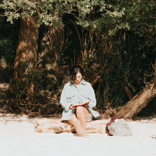 Woman Reads Her Book Sitting On A Wooden Log On The Beach