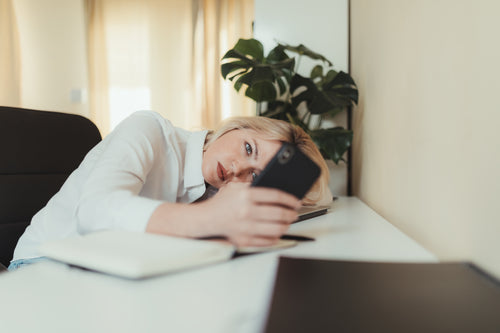 woman rests her head on a table looking at her cellphone