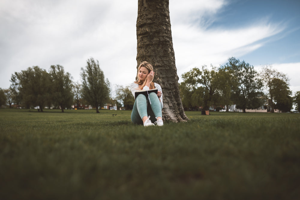 woman sits against a tree trunk and reads a book