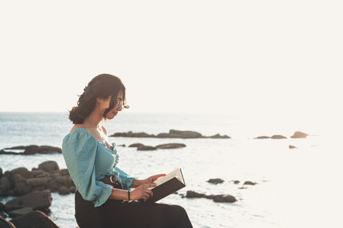 woman sits and reads a book by the rocky shore