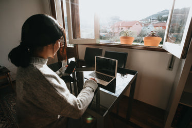 woman sits at near a laptop and looks at her phone