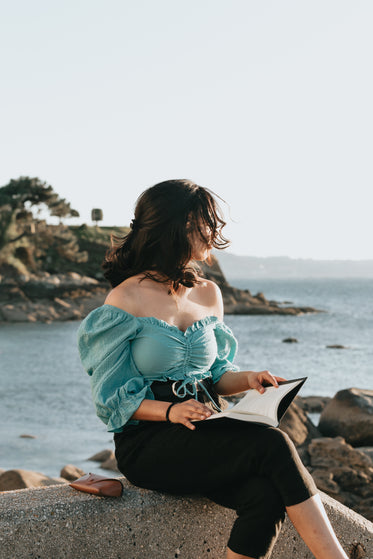 woman sits by the water and holds a novel open