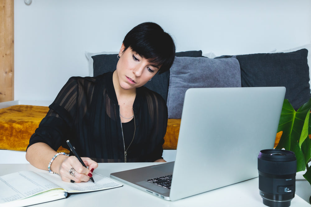 woman sits in front of her laptop and writes in her notebook