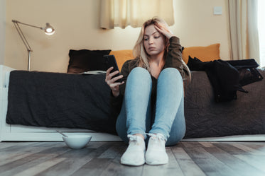 woman sits on floor by her bed looking at her phone