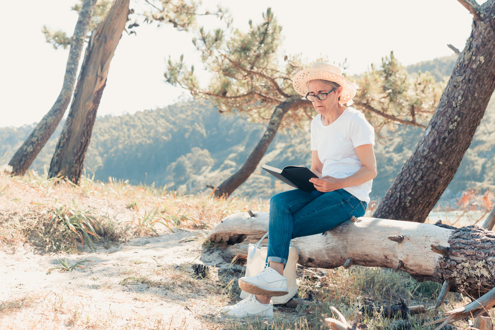 woman sits outdoors and reads a novel on a fallen log