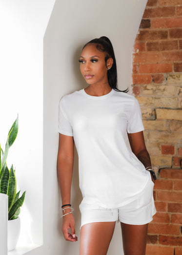 woman wears all white and leans in front of red brick wall