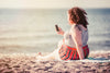 young woman looks at phone while sitting on beach blanket